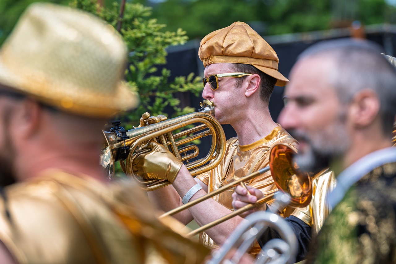Musician dressed in metallic gold with puffed cheeks blowing into a brass flugabone with other players out-of-focus in the foreground